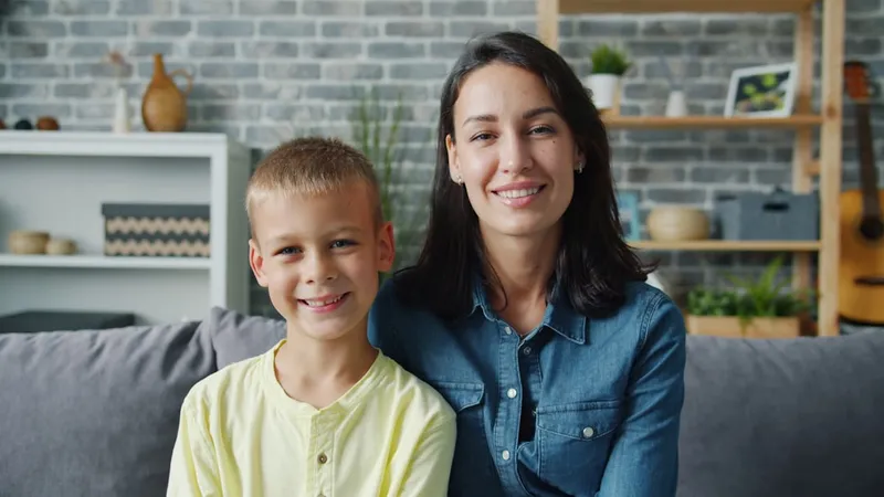 Portrait of happy young family mother and little son smiling sitting together indoors looking at camera. Motherhood, childhood and apartment concept.